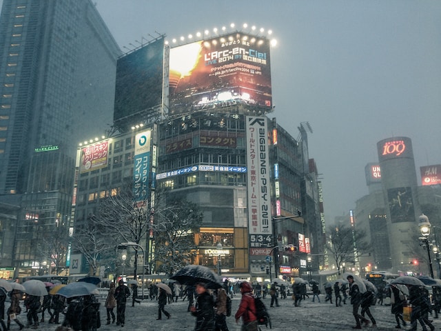 light snow in tokyo. grey skies in shibuya with buildings lit up in neon signs and people walking with umbrellas