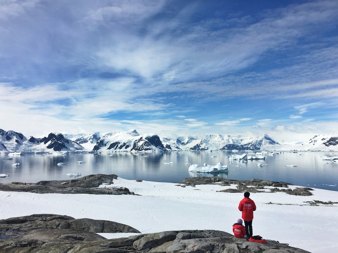 Two people in red parkas sitting on rock in Antarctica, looking out at the ice, open water, and mountainous glaciers ahead