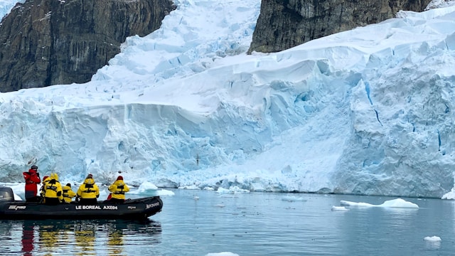 A black zodiac boat carries a group of tourists in yellow jackets with a guide wearing a red jacket. The boat sits in water close to a glacier in Antarctica