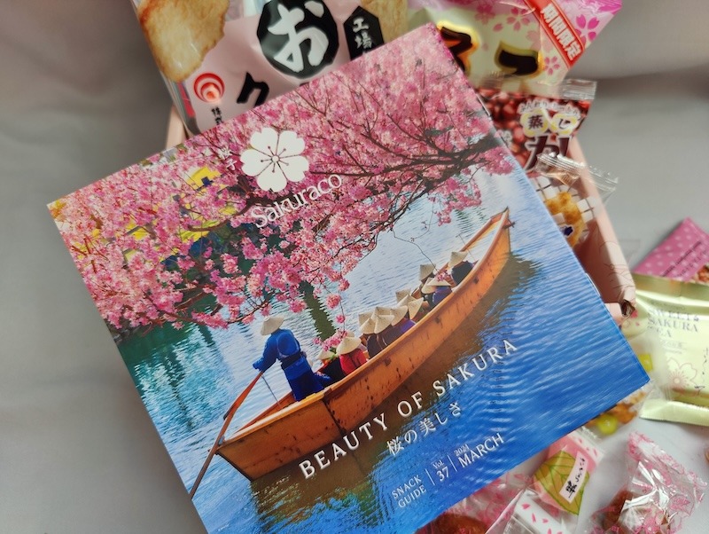 A Sakuraco culture guide cover showing a man wearing a straw hat and traditional robe rowing a group of people in an old wooden canoe under a fully blossomed cherry tree