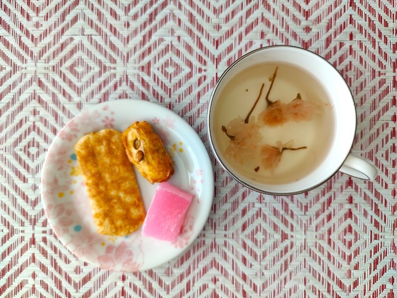 Items opened and staged from a Sakuraco Japanese snack box: two pieces of rick crackers and a pink mochi sit on a floral white and pink ceramic dish next to a cup of sakura tea with three cherry blossoms in the cup