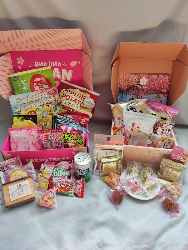 A photo showing Sakuraco vs. Tokyo Treat boxes side by side. On the left is a Tokyo Treat box, opened, and showing the contents: matcha latte KitKat, potato chips, sakura rice crackers and cookies, castella cake, candy, and a Japanese soda. On the right is the Sakuraco box, opened, showing the contents: traditional rice crackers, tea, sweets, baked goods, mochi, and a souvenir ceramic dish