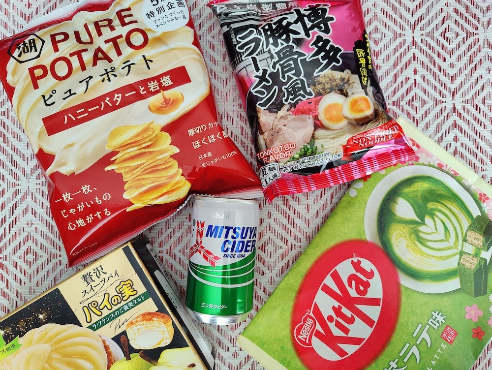 Flatlay of some snacks from a Tokyo Treat snack box: potato chips, Hakata tonkatsu ramen, matcha latte KitKat, Mitsuya cider, and mini pear tart
