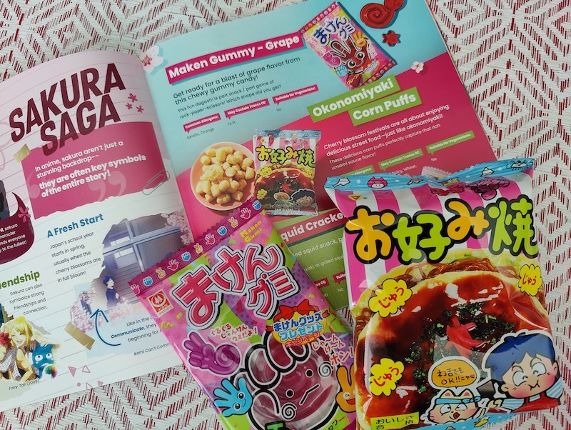 A Tokyo Treat culture guide opened to show a page about the grape gummy candy and okonomiyaki corn puff snack included in the box. Below the culture guide are the actual two snack items