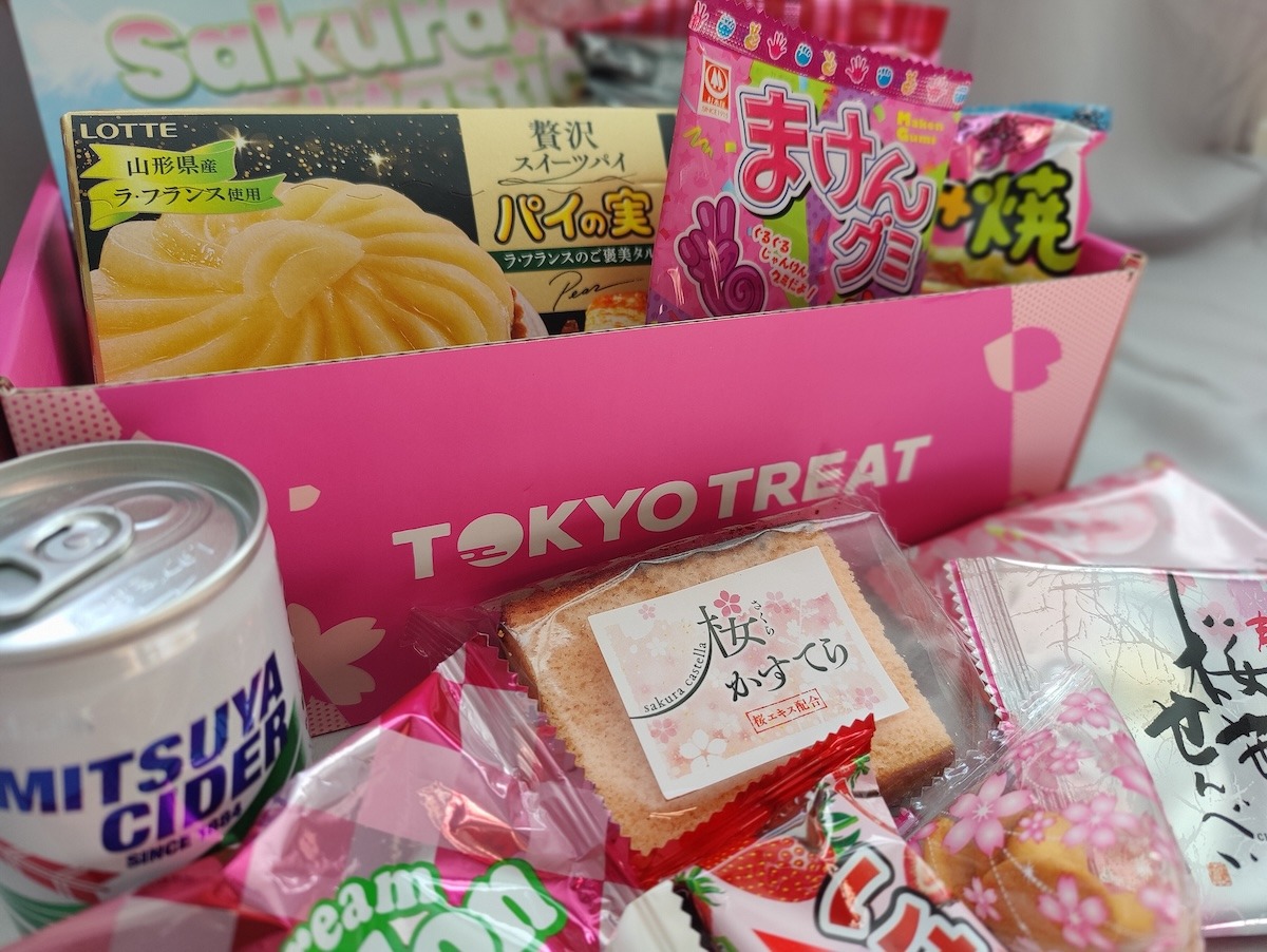 A close-up shot of the snacks for this Tokyo Treat review. The pink box is opened with the snacks surrounding it: pear tart, kids grape gummy candy, Mitsuya Cider, and sakura-flavored cookies, cake, and rice cracker