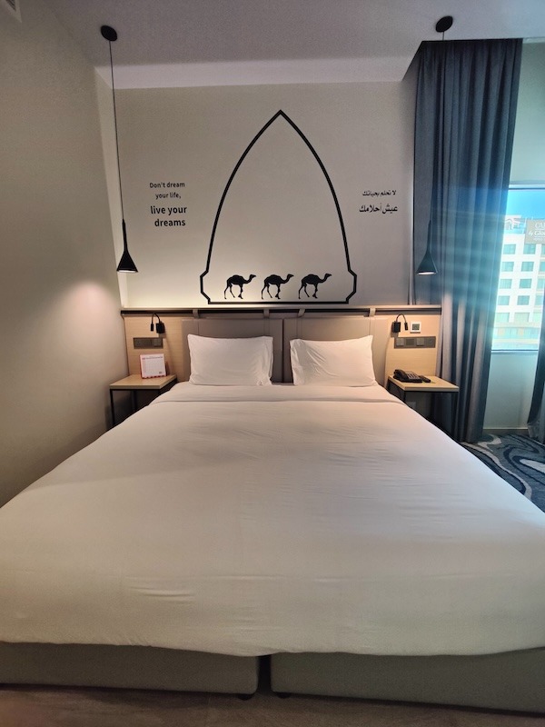 A bed at the Swiss-Belinn Airport Muscat hotel. The bed has white bedsheets with two nightstands, one on each side of the bed. Above the bed is a simple silhouette of three camels along with the phrase: "don't dream your life, live your dreams" on the left and the phrase in Arabic on the right.