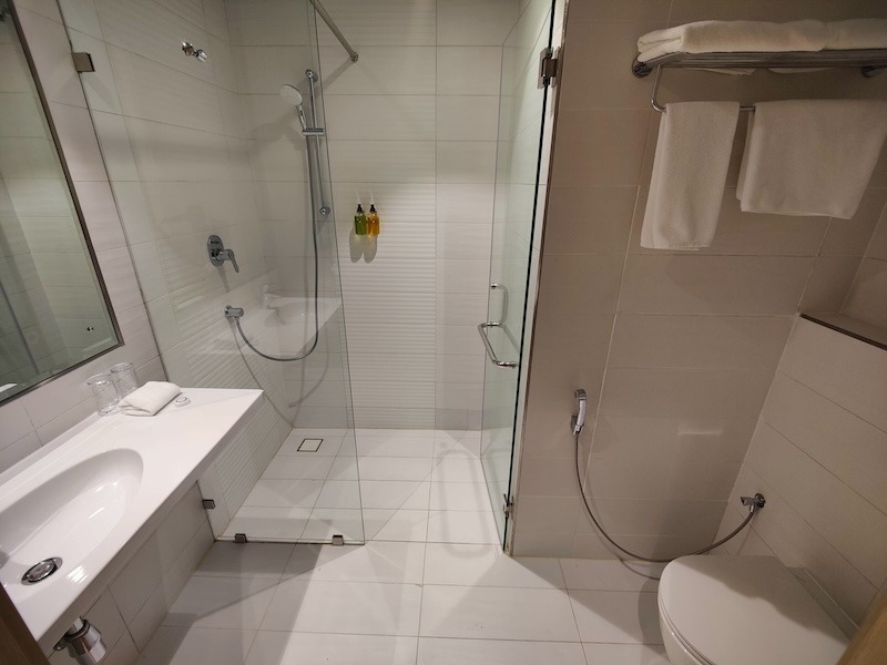 The bathroom inside a standard room at the Swiss-Belinn Airport Muscat hotel. On the left is a sink and long counter, all in white. The shower is in the venter with a glass door. To the right is the toilet and bidet along with towels on the racks above.