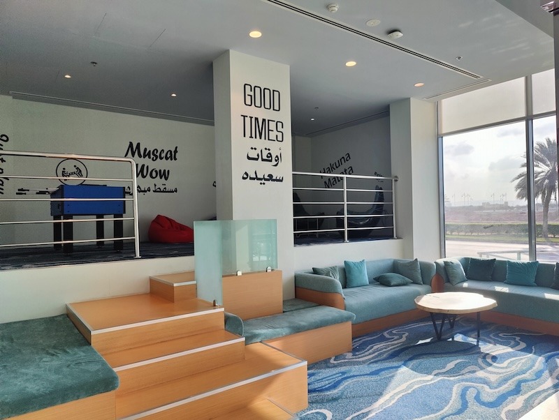 The lounge area at the Swiss-Belinn Airport Muscat. It is two-tiered with a few steps separating the levels. The bottom level has couches. The upper level has bean bag chairs and a fooseball table. There is a pillar with the words "good times" in English and Arabic.