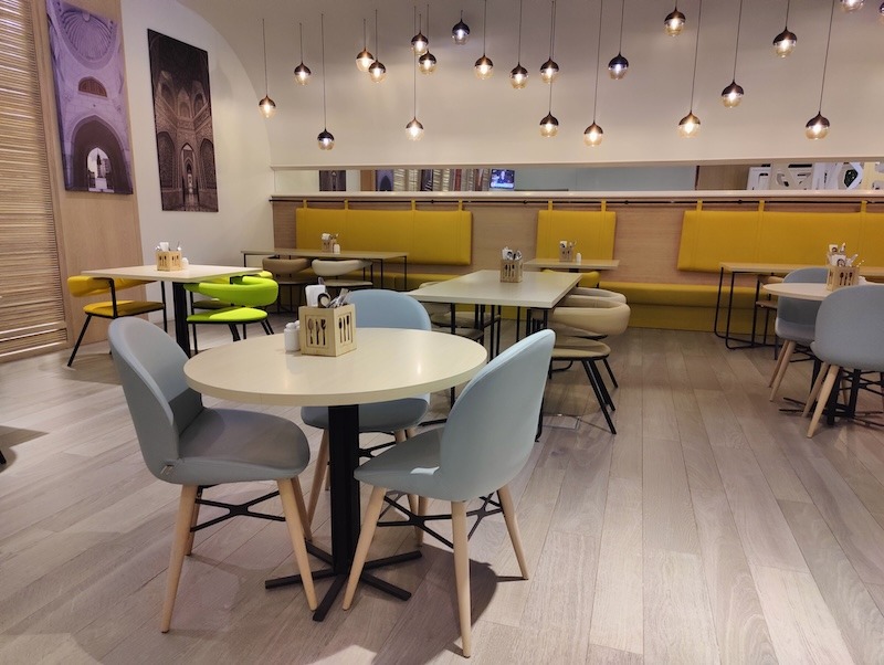 The dining area at the Swiss Belinn Airport Muscat Oman. This is also called the Swiss Cafe and includes small circular and round tables with a variety of cushioned, Ikea-looking chairs. On the far tables are also booths with yellow cushioning. Individual artsy lamps hang from the ceiling.