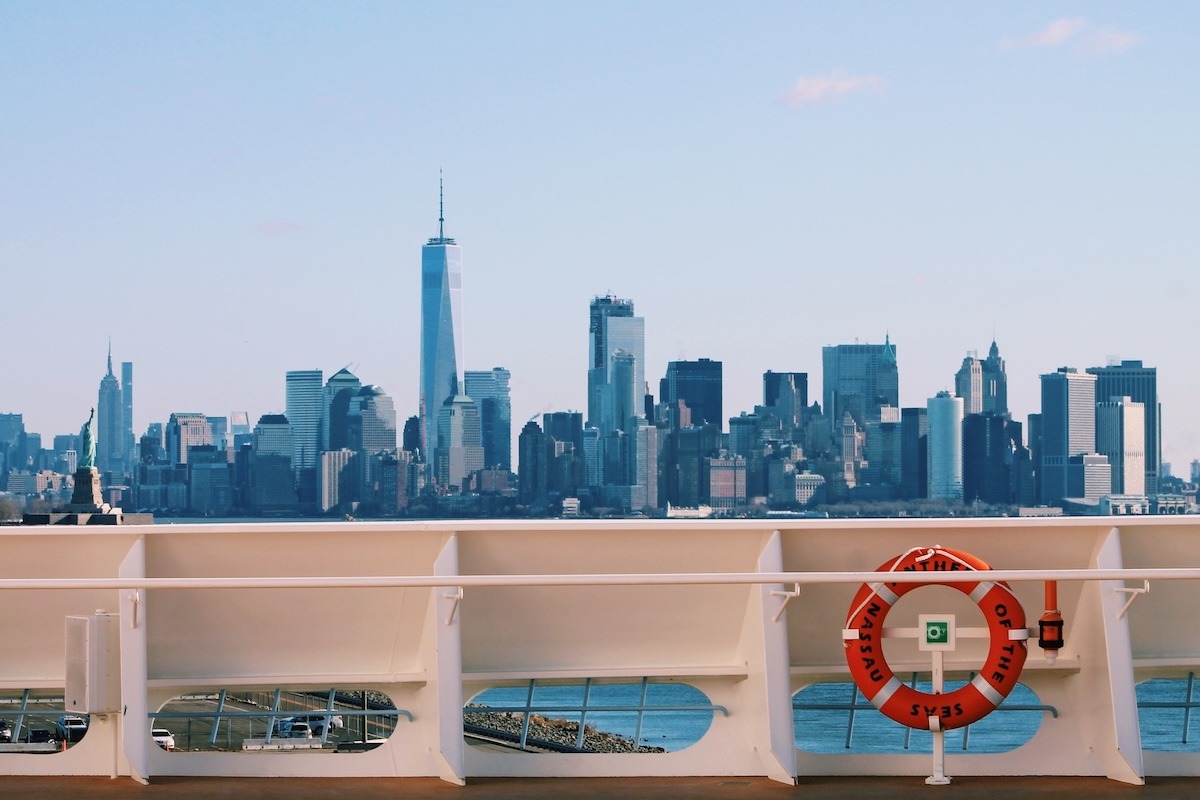 Anthem of the Seas docked in front of the New York City skyline. In the foreground: railing of the ship and orange life ring. Background: Statue of Liberty, Empire State Building, One World Trade Center, and other buildings.