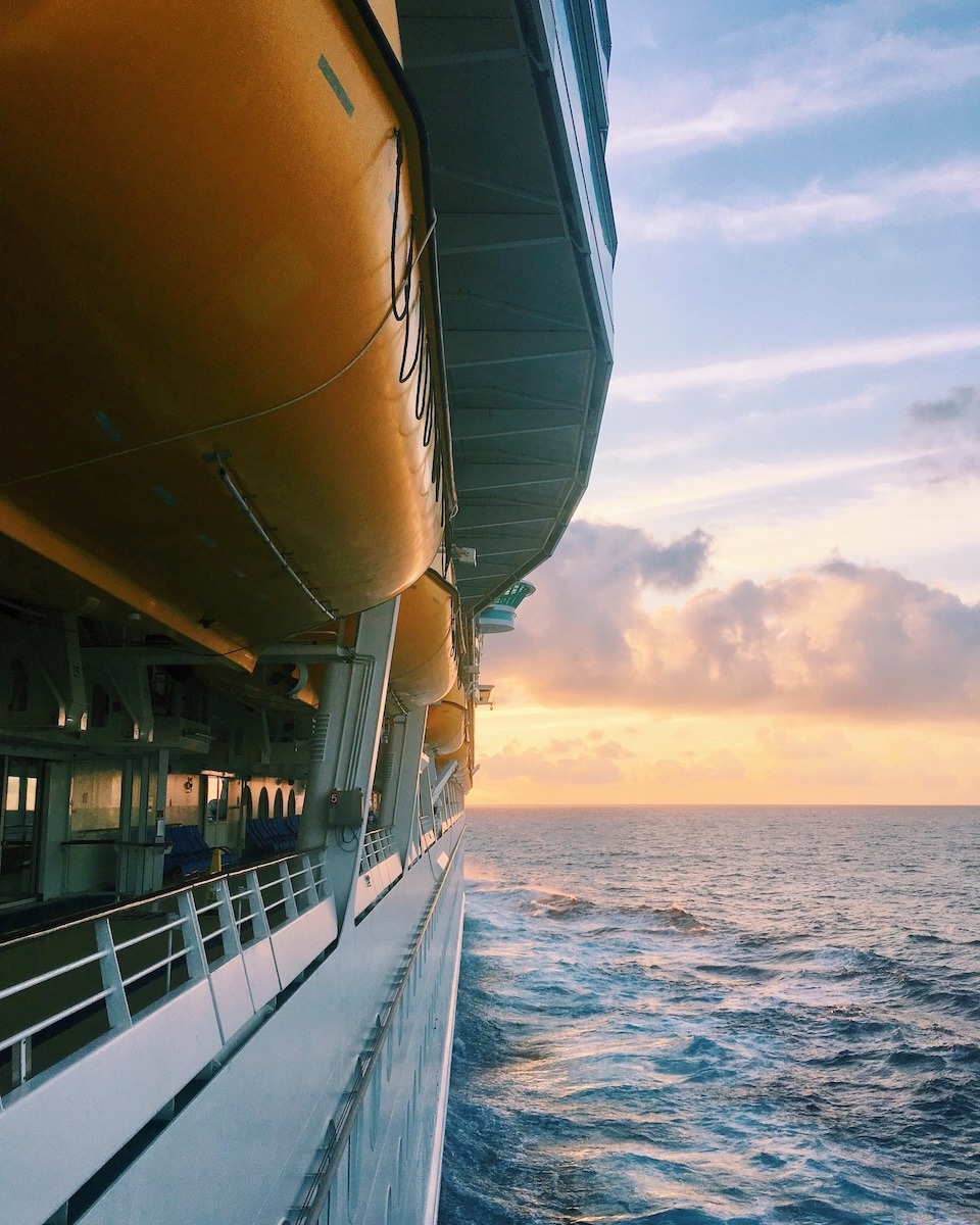 A photo off the side of a ship. On the left is the exterior of the Freedom of the Seas with lifeboats above. To the right is a sunset and calm waters.