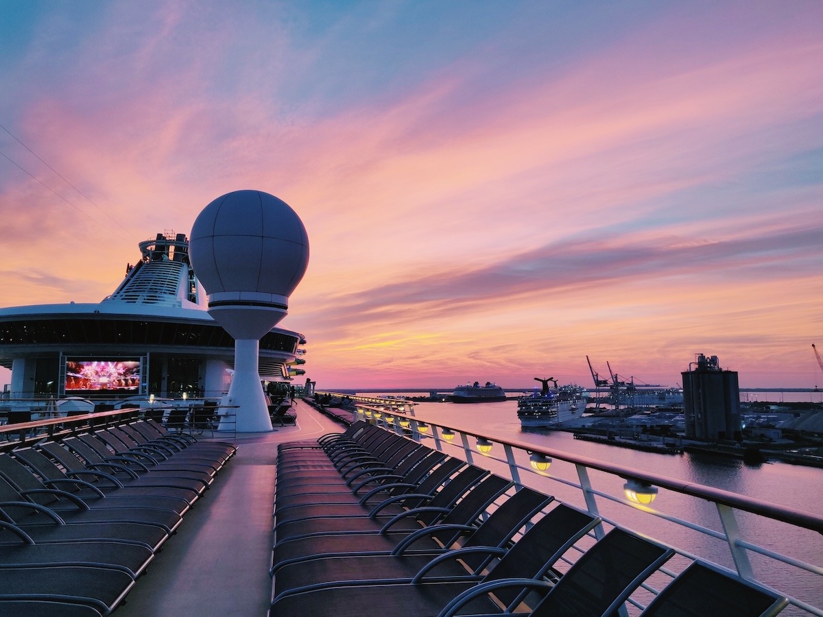 A sunset photo on the top deck of the Mariner of the Seas in port. The photo of the deck incudes empty lounge chairs under a pink and orange sunset with two other ships docked in port to the right.