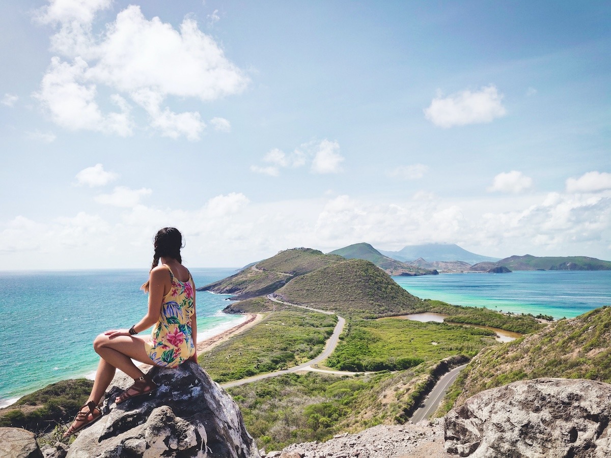 A woman in a floral yellow romper sits on a rock at Timothy Hill in St. Kitts overlooking the Atlantic Ocean and Caribbean Sea. Between the waters are small green hills separating the two.
