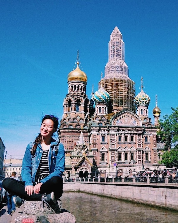 Michelle sits in front of The Church of the Savior on Spilled Blood in St. Petersburg. She has a side braid and is wearing a blue denim jacket, black and white striped shirt, black jeans, and gray tennis shoes