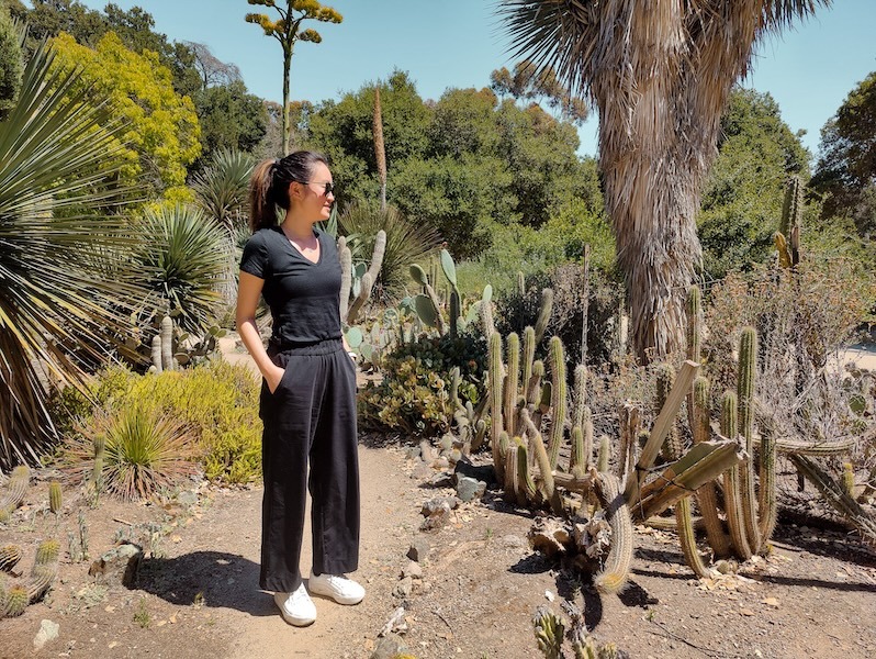 A woman stands in a cactus garden surround by cacti and trees in the background. She is wearing Unbound Merino's v-neck t-shirt in black, the lightweight travel pants in black, and white sneakers