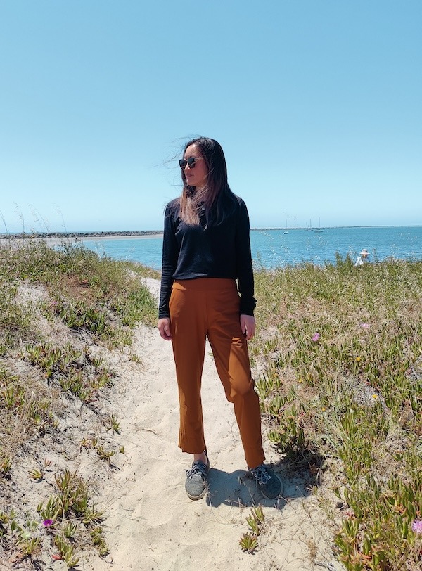 A woman stands at the beach on a path with grass on the sides and the ocean in the background. She is wearing sunglasses, an Unbound Merino long sleeve crew shirt in black, pecan pants, and gray sneakers