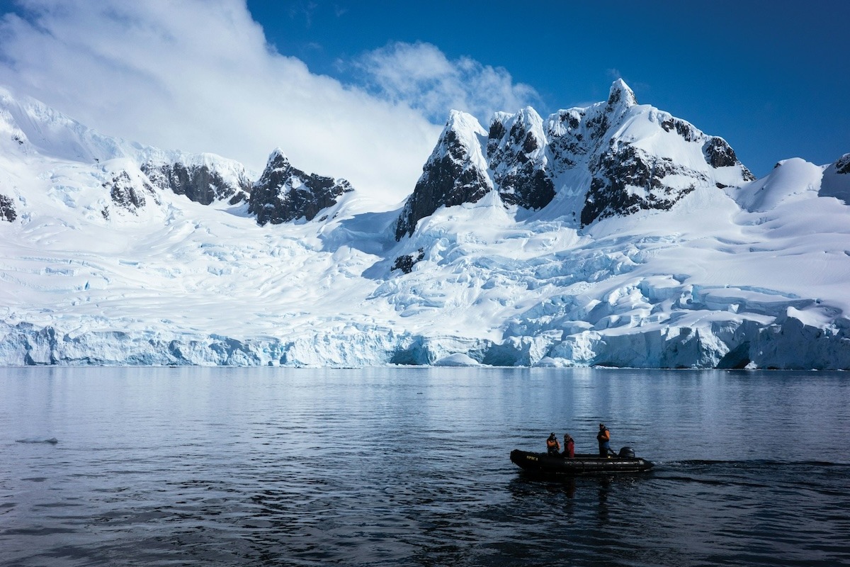 A zodiac with three visitors in red parks sit in the water looking at the frozen glacial mountain in the background in Antarctica