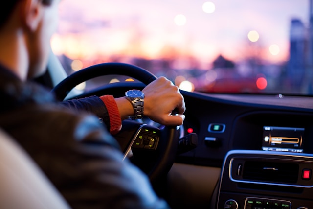 A man drives a car. His left hand is on the steering wheel