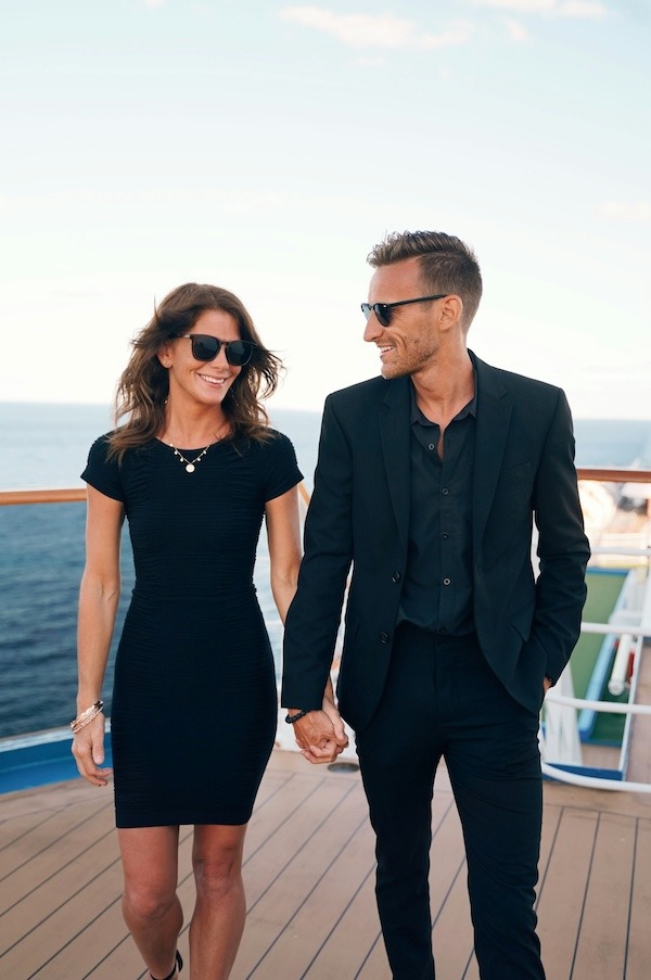 A couple holds hands on the deck of a ship. The woman wears a short sleeve black mini dress. The man wears a dark navy suit.