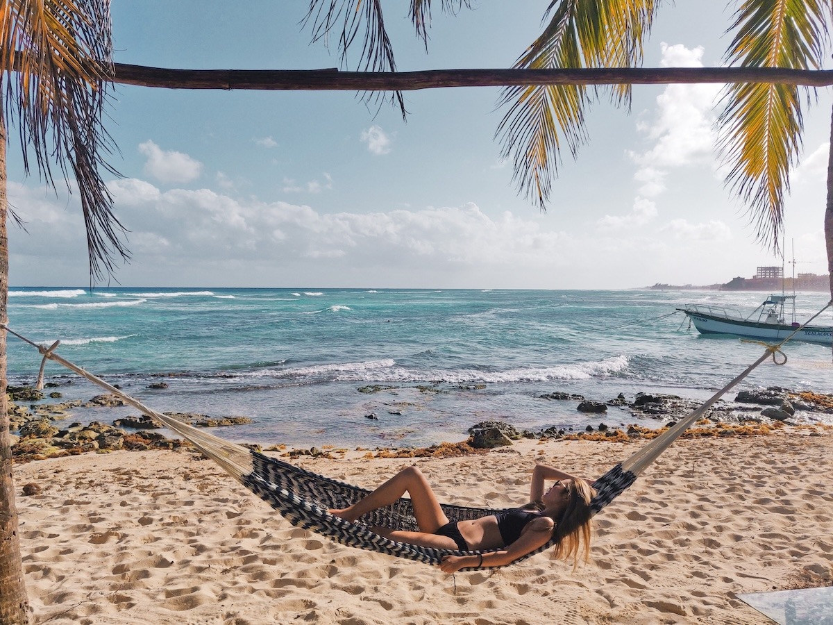 A girl in a black bikini lays in a hammock under palm trees on a beach, looking at the sea