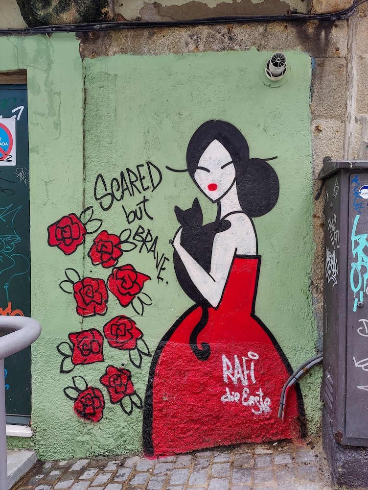 Street art of a woman in a red dress holding a black cat with the text "scared but brave"