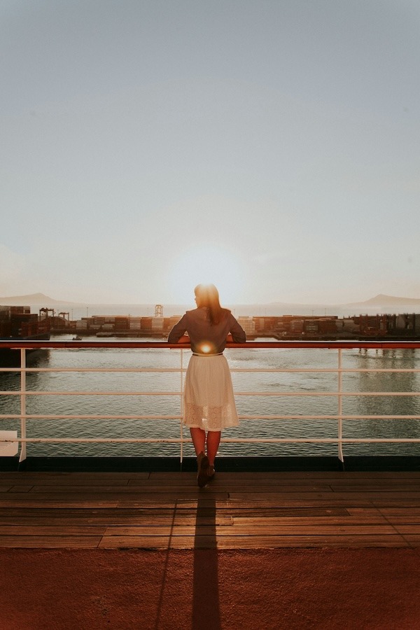 A woman stands on the deck of a ship looking out at the water. She wears a white dress.