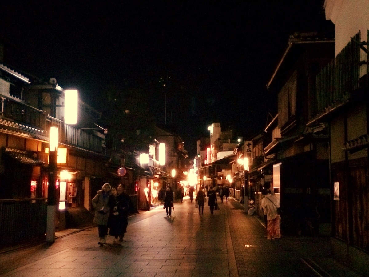 Winter in Kyoto during nighttime. Traditional houses are dimly lit along a pedestrian street as people in coats walk through