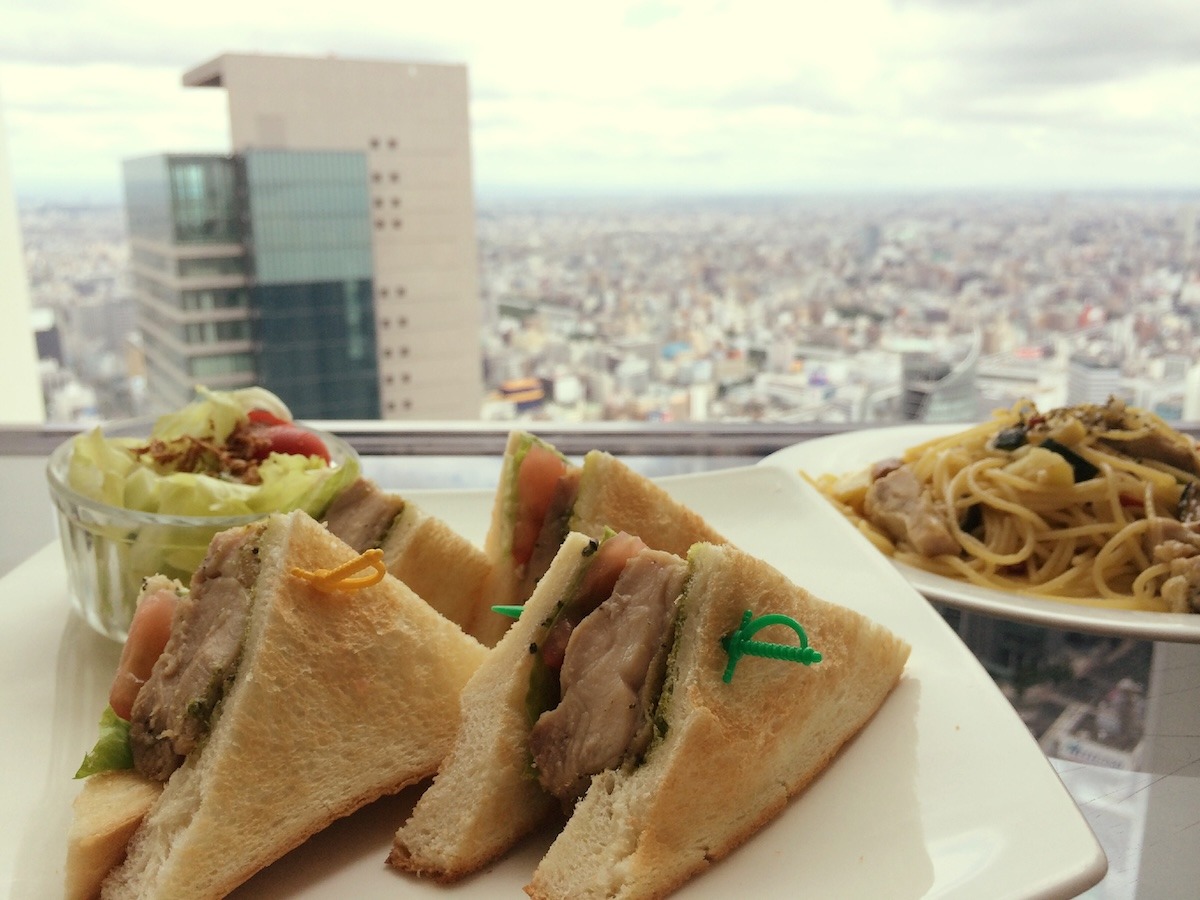 A sandwich sliced into triangular quarters with a small salad and a plate of pasta sit in front of the Nagoya skyline at Cafe Du Ciel