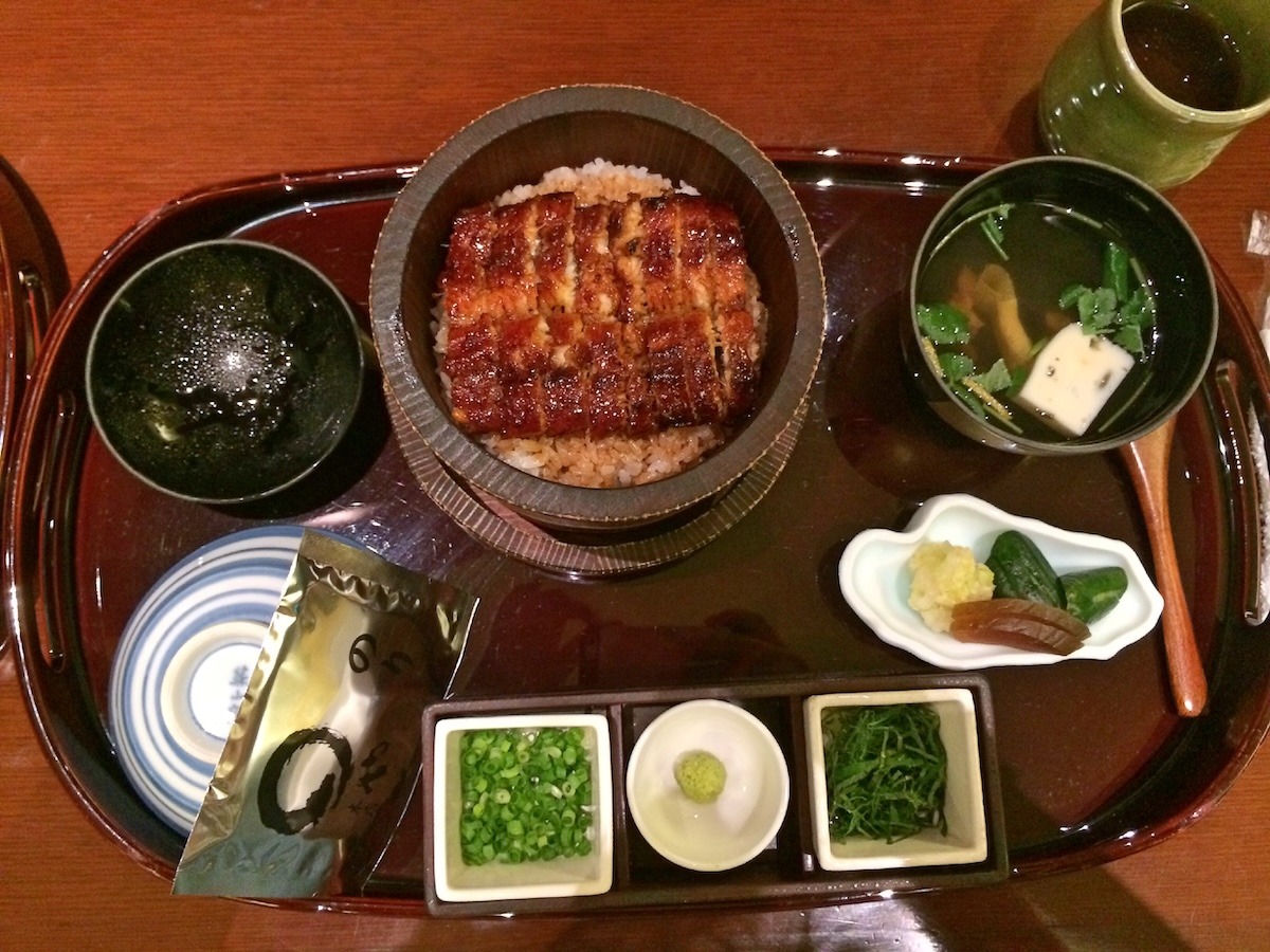 A hitsumabushi (barbecued eel over rice) meal set. The bowl of rice with eel sits on a tray along with a cup of soup, pickles, green onion garnish, and tea