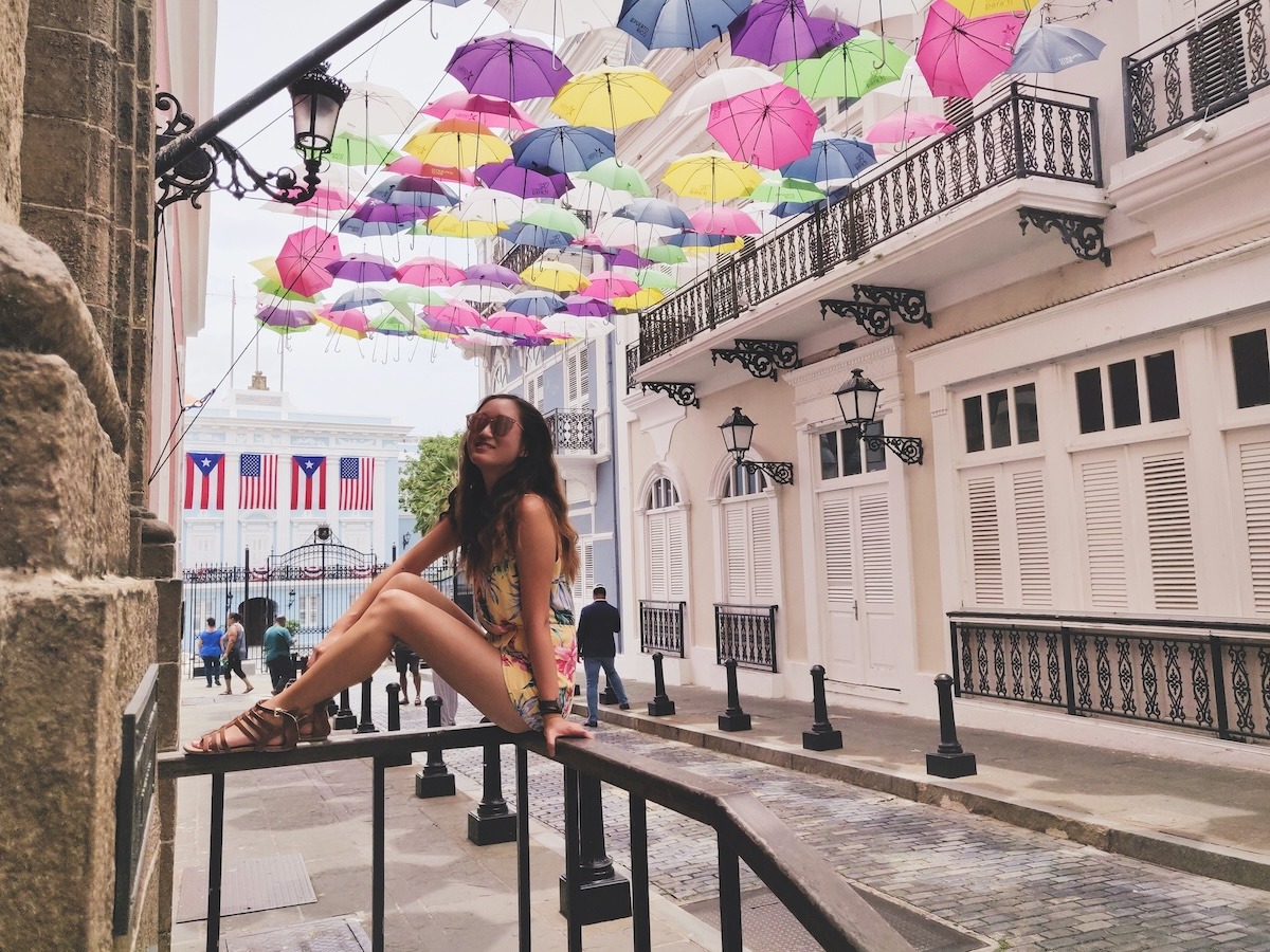 A girl in a yellow floral romper sits on a railing under colorful umbrellas on San Juan Puerto Rico's famous Umbrella Street. Puerto Rican and American flags hang in the background