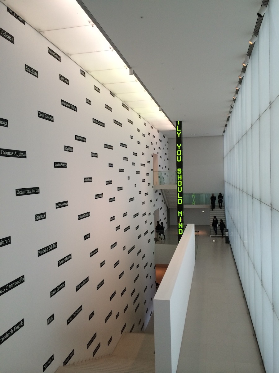 An interior stairway of the Nagoya City Art Museum. The walls are white and names are printed on black rectangles along the wall. In the background there is a floor-to-ceiling vertical LED sign with green letters ("[cut off]ly you should mind" appear in the picture)