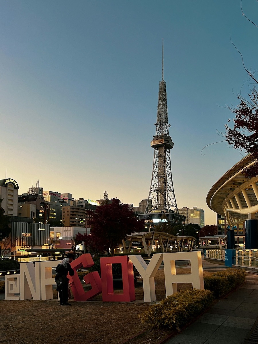 large letters "NAGOYA" sit in front of the Nagoya TV tower and Oasis 21 aquaship at dusk