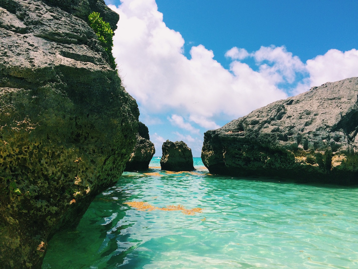 A small cove tucked away near a beach in Bermuda; pictured are still, aquamarine waters with three large rocks forming a small cove