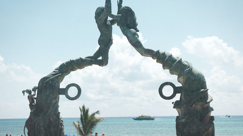 two large mermaid statues joining hands to form a gateway in front of the ocean; tourists walking in front of it