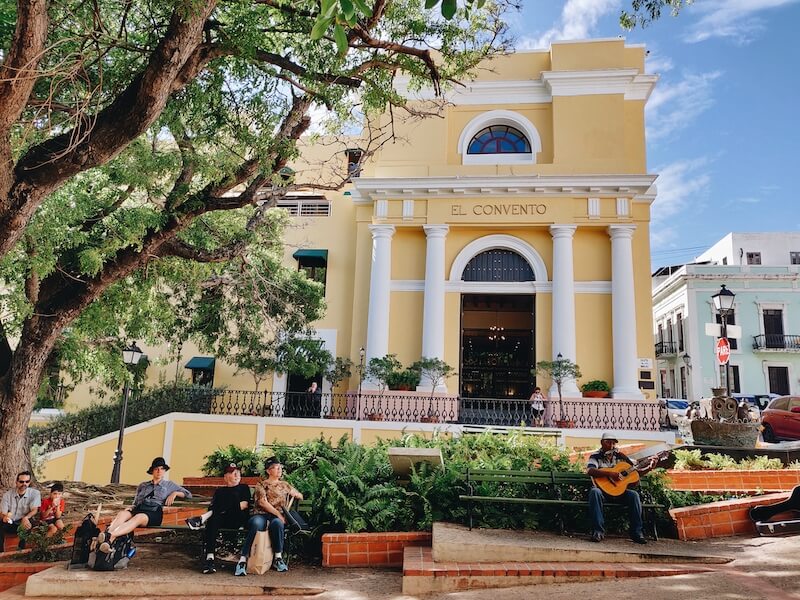 Bright yellow hotel El Convento with large trees, and locals sitting outside listening to a man playing the guitar