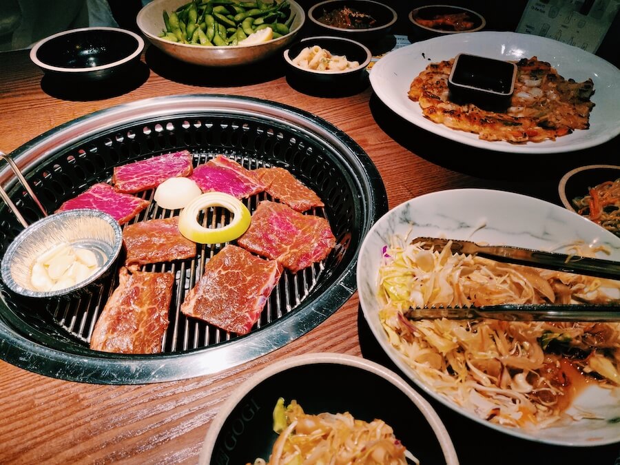 Picture of slices of beef, onions, and garlic on the grill. Also pictured: edamame, seafood pancake, and cabbage salad
