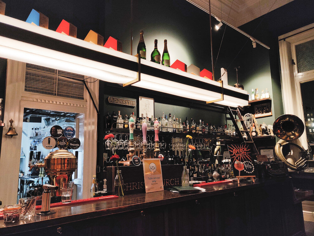 Interior bar inside OGB Bar. Three bottles of champagne on the top shelf, bottles of liquor at eye-level, and a dark wooden bar with brass hardware