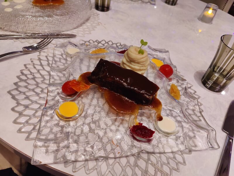 a piece of short rib sits on a transparent square platter with a side of mashed potatoes. the platter has divots for various colorful sauces