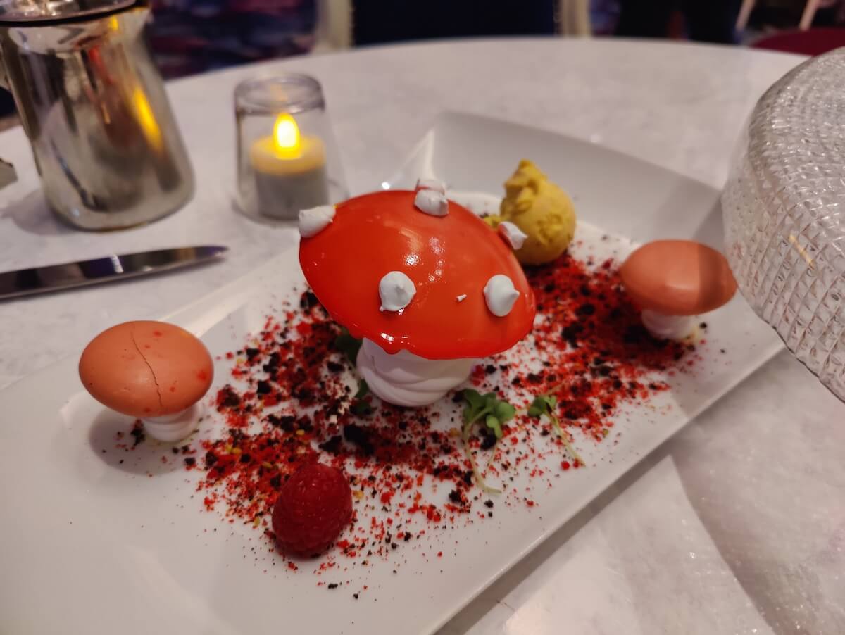 a red-topped mushroom with white dots frosted on top sits on a plate with two other tan-topped mushrooms on a white plate with red and black crumbs.