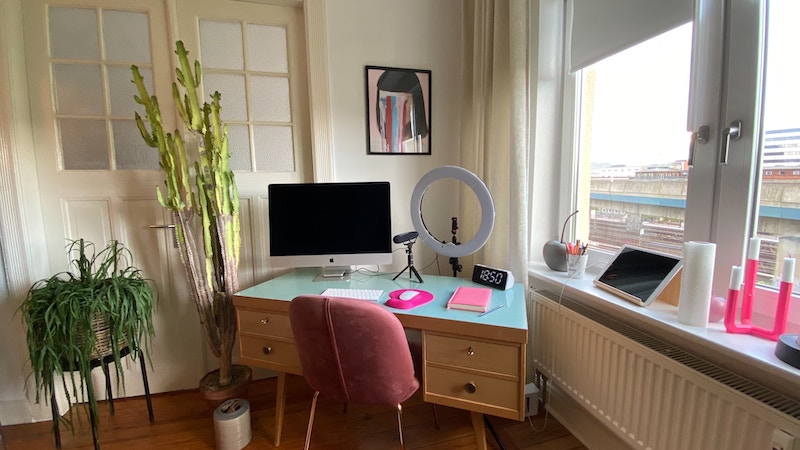 The corner of a room set up for a content creator, one of the best jobs that travel with no experience. There is a desk with a computer monitor, keyboard, mouse, webcam, and ringlight.