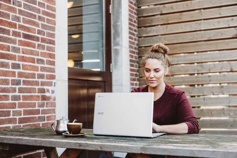 A woman wearing a red top sits at a wooden table outside working on a laptop with a hot beverage