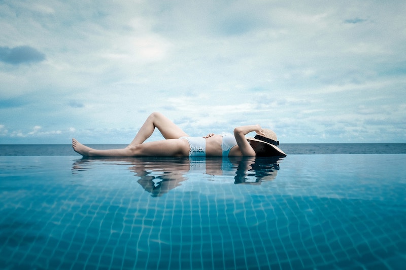 A woman in a pale blue two piece bathing suit sits at the edge of an infinity pool with her face covered with a straw hat