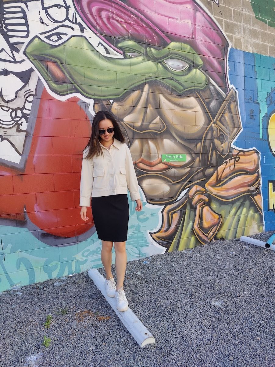 A woman poses in front of a graffiti wall. She wears a white lightweight jacket and black dress underneath