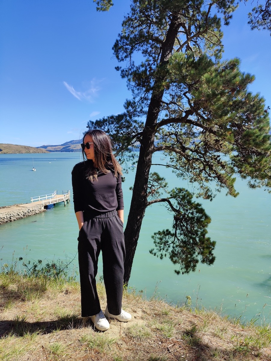 A woman stands in front of a blue lake. She wears a black long sleeve shirt and black pants