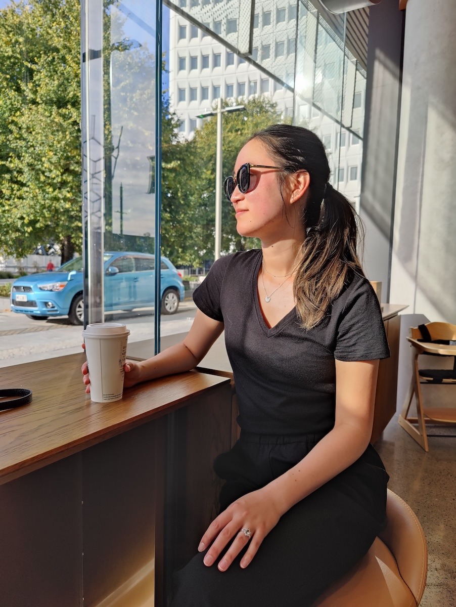 A woman wearing a black tshirt and pants sits at a cafe holding a cup of coffee while looking out the window