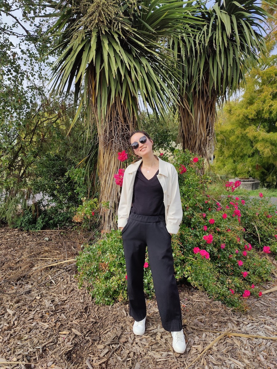 A woman wears a white lightweight jacket over a black vneck tshirt and black pants. She stands in a garden with woodchips on the ground and flowers and trees in the background