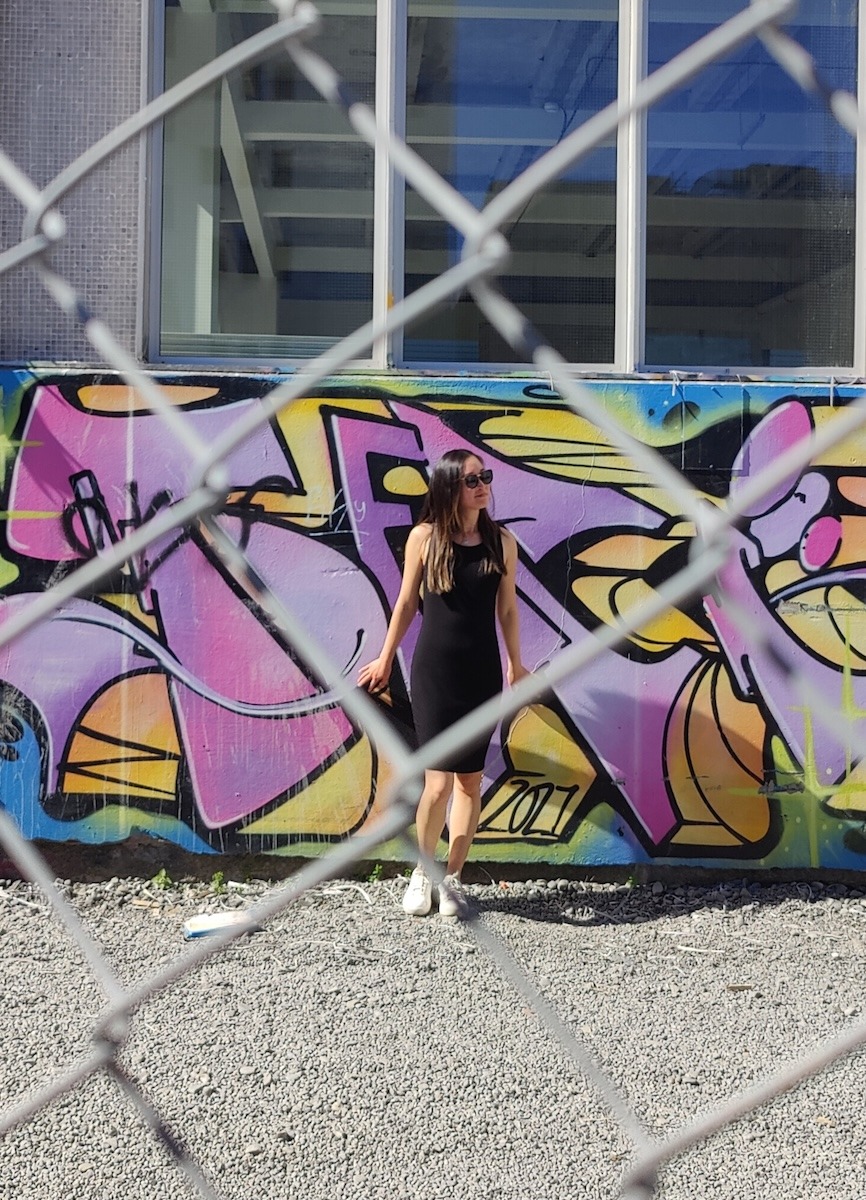 An artsy photo looking through a fence toward a building with graffiti. A woman wearing a black mini dress stands in front of the graffiti