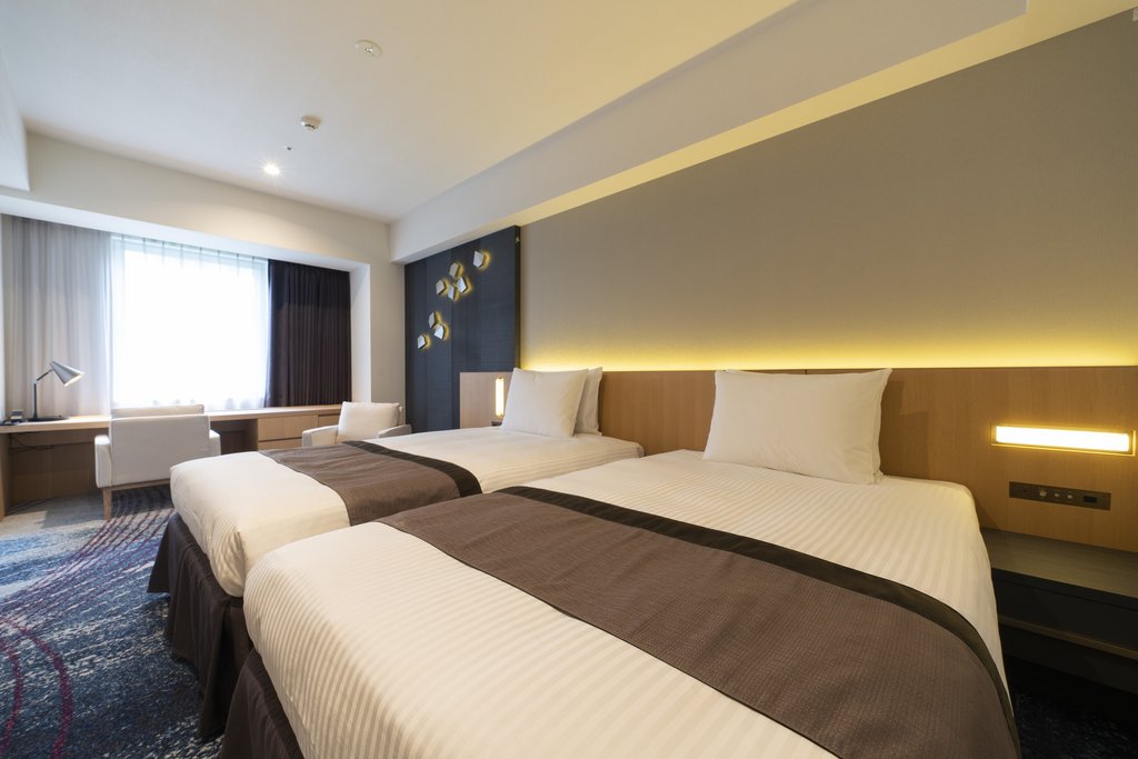 Two beds in a hotel room with warm back lighting. There's a desk with a chair and lamp on the left in front of a window.