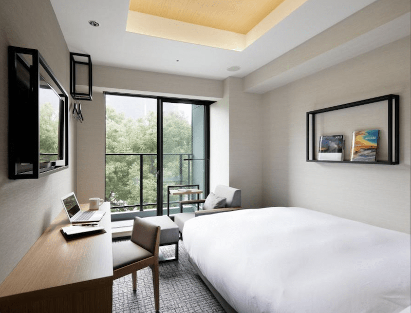 A modern hotel room featuring a comfortable bed, a wooden desk with a laptop, and a cozy chair. The large window offers a view of greenery outside, with natural light illuminating the space.