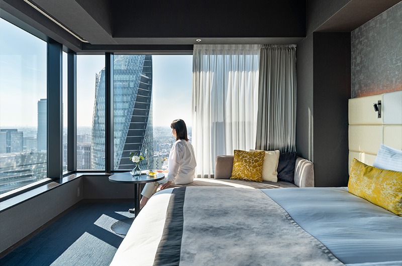 A woman sitting on a bed in a modern hotel room, enjoying breakfast with a view of the city skyline through large windows.